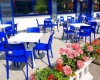 Terasa kaviarne s modrými stoličkami a bielymi stolmi vedľa kvetináča s ružovými kvetmi.