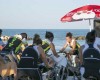 Ľudia cvičia na stacionárnych bicykloch vonku pri mori pod slnečníkom.