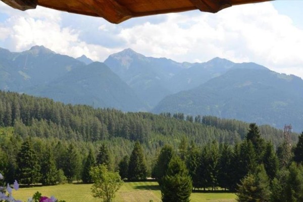 Zalesnená krajina s horským pohorím v pozadí a dreveným okrajom strechy hore.