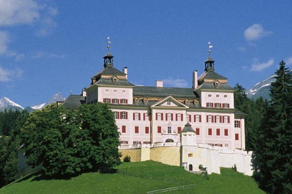 Historický zámok s vežičkami obklopený zeleňou a horami.