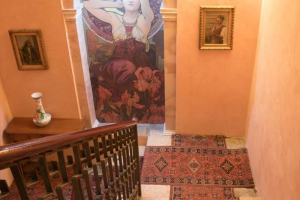 Schodisko s kobercovými behúňmi, ozdobnými obrazmi na stenách a veľkou art nouveau maľbou postavy.