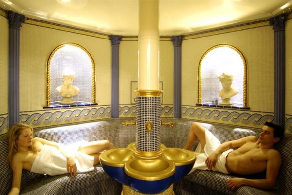 Dvaja ľudia relaxujú na lehátkach v luxusnej miestnosti s mozaikovou výzdobou.