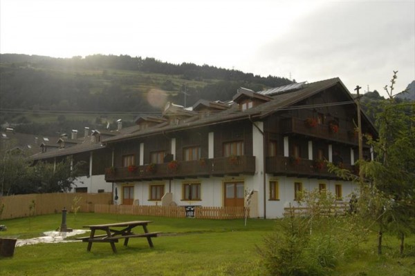 Penzión s drevenými balkónmi a lavičkou v popredí na pozadí kopcov.