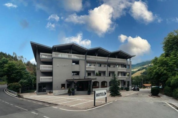 Moderný šedý hotel s balkónmi a šikmou strechou obklopený prírodou.