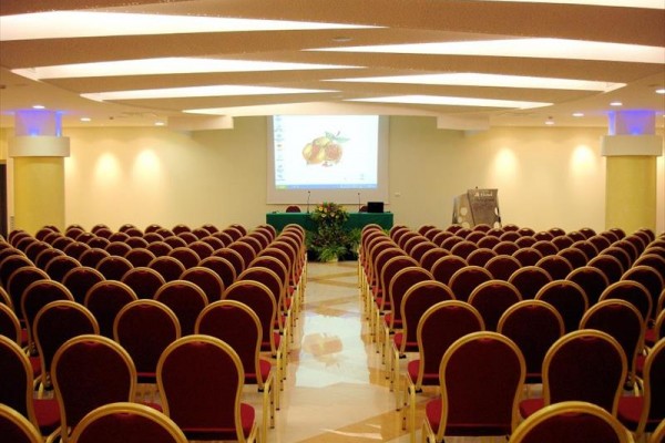 Konferenčná sála s prázdnymi radmi červených stoličiek a premietacím plátnom na pódiu.