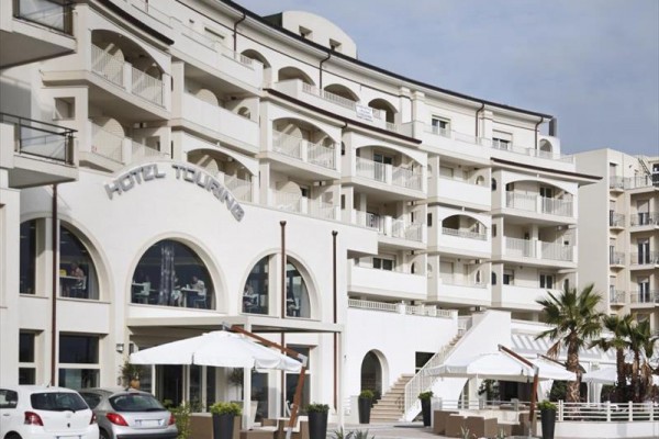 Moderný hotel so svetlou fasádou a vonkajšou terasou s bielymi slnečníkmi.