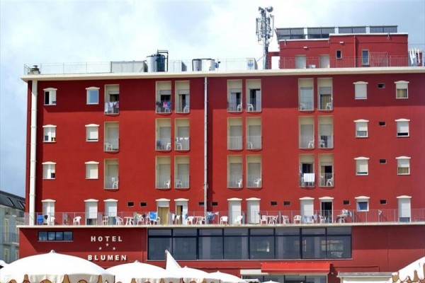 Tehlovo červená fasáda hotela s balkónmi a vonkajším posedením.