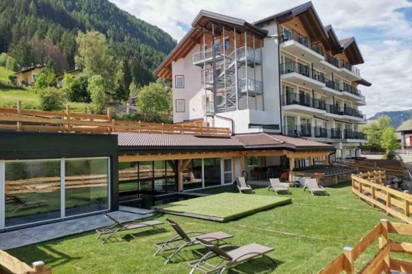 Moderný hotel s terasou a lehátkami na sviežej zelenej záhrade obklopený horami.