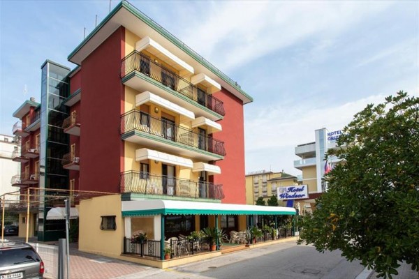 Štvorposchodový hotel s verandou a balkónmi, obklopený zeleňou a susednými budovami.