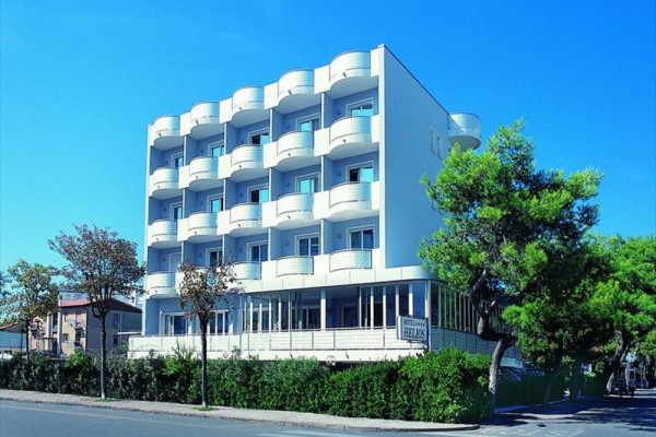 Štvorposchodový biely hotel s balkónmi, obklopený stromami a kríkmi, pod modrou oblohou.