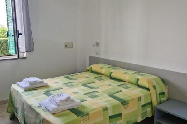 Spálňa s otvoreným oknom, manželskou posteľou so zelenožltým prehozom a zloženými uterákmi.
