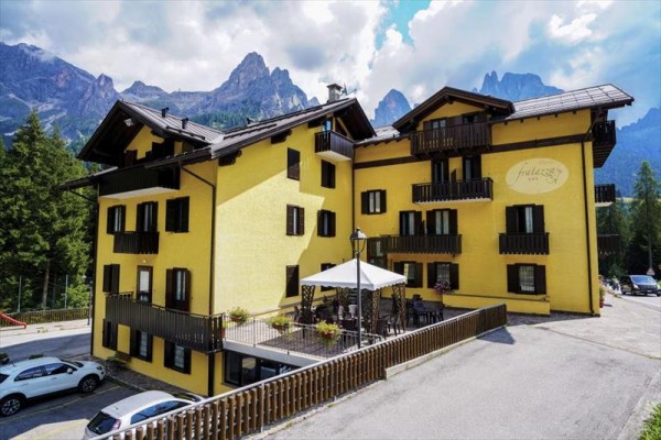 Žltý hotel s drevenými balkónmi obklopený horami a lesmi.
