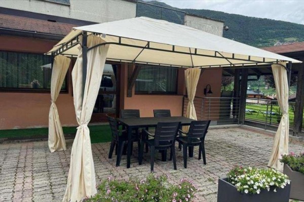 Stôl so šiestimi stoličkami pod pergolou s bielou plachtou na terase, obklopený kvetmi.