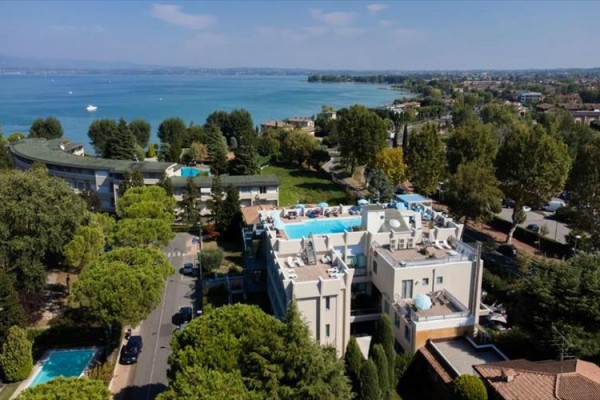 Pohľad na hotel s bazénom na streche pri jazere obklopený stromami a ďalšími budovami.  