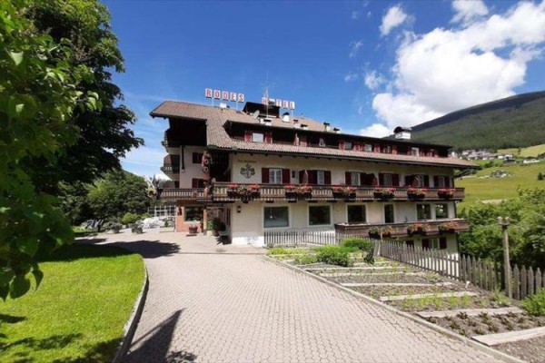 Hotel s ozdobnými balkónmi obklopený zeleňou a kopcami počas slnečného dňa.