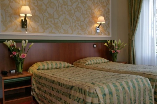 Dve jednolôžkové postele s kvetmi na nočných stolíkoch v útulnej izbe s elegantnou tapetou a nástennými lampami.  