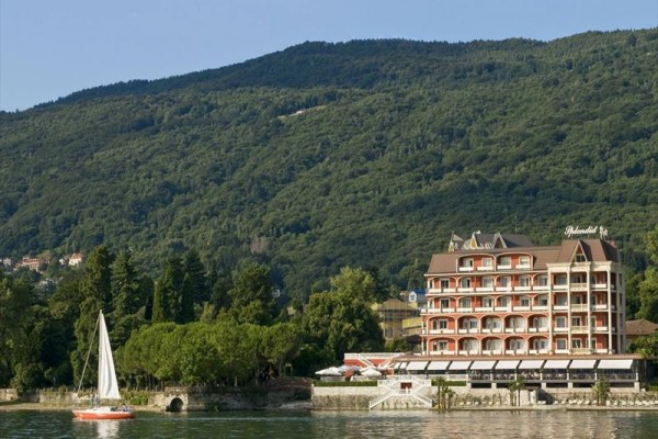 Hotel pri jazere s plachetnicou v popredí a zalesnenými kopcami v pozadí.