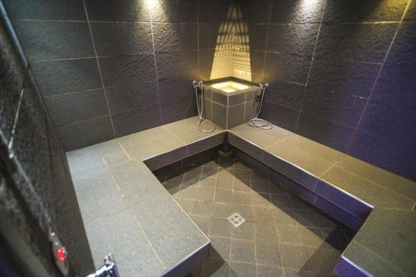 Parná sauna s lavicami a sprchovými hlavicami na tmavej dlažbe. 