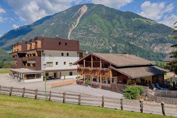 Horský hotel s drevenými balkónmi a kvetinami pred zeleným kopcom a modrou oblohou.  