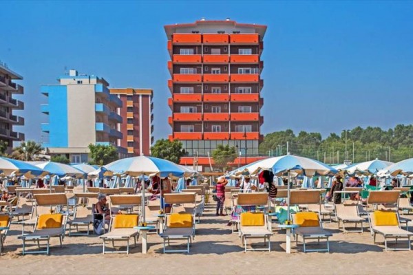 Piesočná pláž s ležadlami a slnečníkmi pred moderným hotelom.