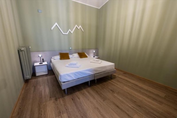 Spálňa s dvojposteľou, bielymi nočnými stolíkmi a dekoráciou v tvare hôr na stene.