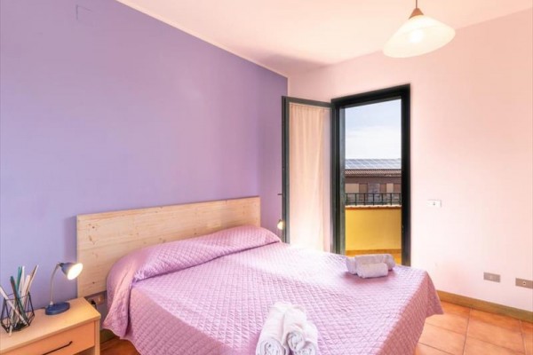 Spálňa s fialovou stenou, manželskou posteľou s ružovým prehozom a balkónovými dverami.