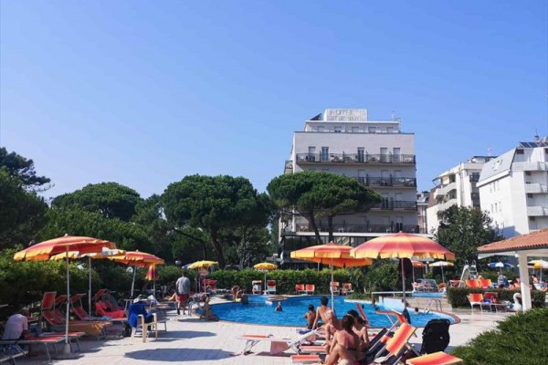 Ľudia relaxujú pri bazéne s lehátkami a slnečníkmi, v pozadí sú stromy a hotel.