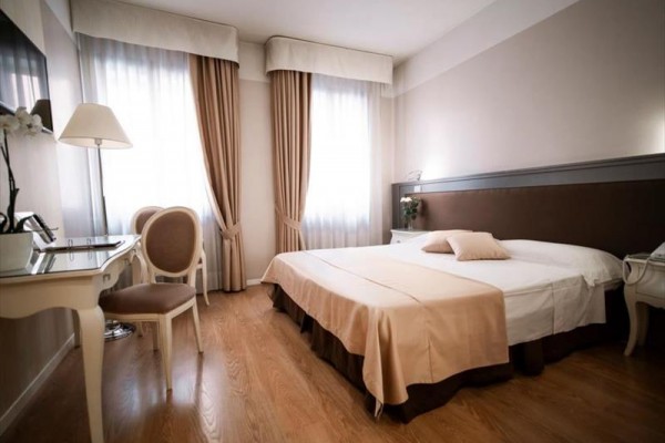 Elegantná hotelová izba s manželskou posteľou a nábytkom v krémových odtieňoch.