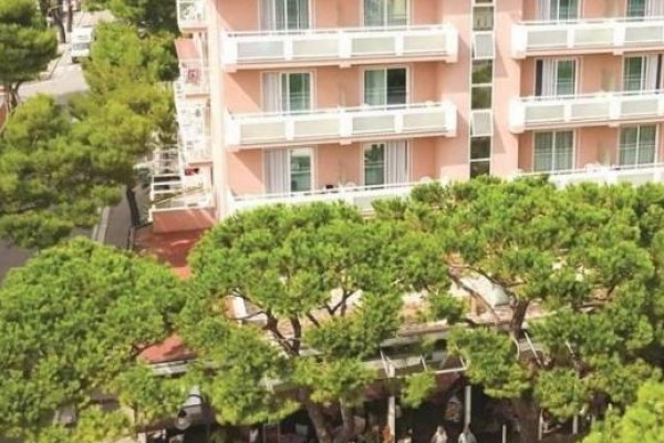 Ružová budova s balkónmi obklopená vysokými stromami.