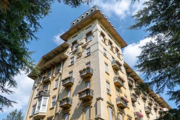 Historická budova hotela s balkónmi a ozdobnými detailami pod modrou oblohou medzi stromami.  