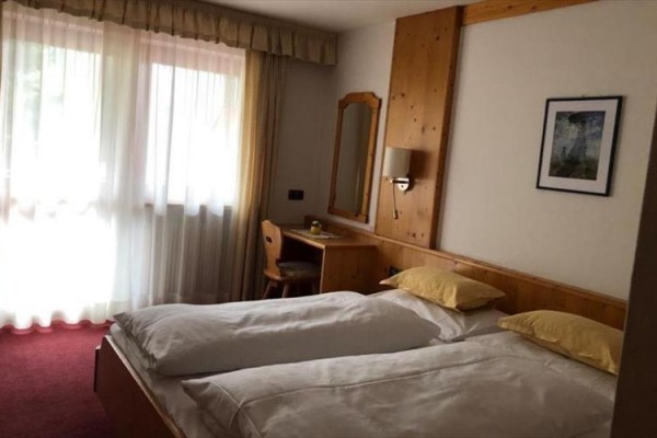 Dvojposteľová izba s bielou posteľnou bielizňou, dreveným nábytkom a oknom so záclonami.  
