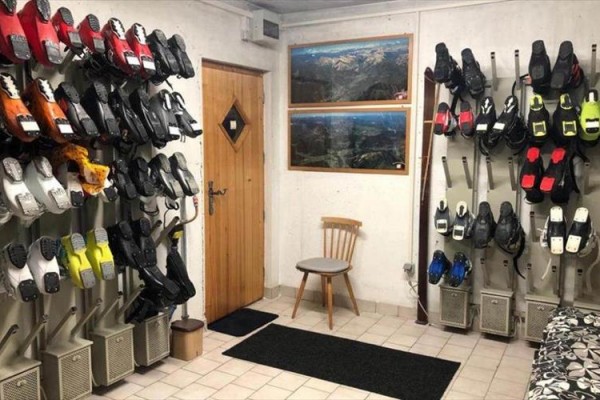 Miestnosť na sušenie lyžiarok s radmi topánok na sušiakoch a drevenou stoličkou.