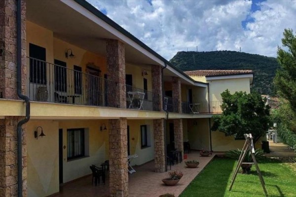 Dvojpodlažná budova s terasami, stoličkami a trávnatým dvorom na pozadí kopcov.