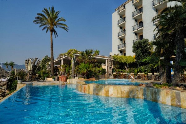 Hotel s bazénom obklopený palmami a rastlinami pod modrou oblohou.