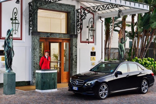 Čierny Mercedes zaparkovaný pred vstupom do luxusného hotela s vrátnikom vo dverách v červenej uniforme.  