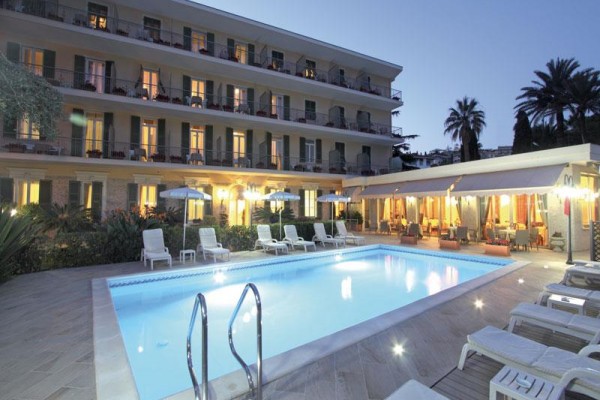 Večerný pohľad na osvetlený bazén pred trojposchodovým hotelom s palmami v pozadí.