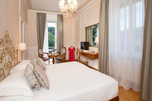 Elegantná hotelová izba s veľkou posteľou, kreslami a výhľadom na zeleň.