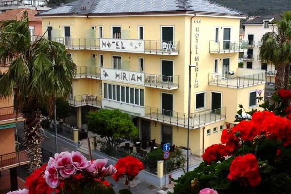 Žltý trojpodlažný hotel s balkónmi a kvetmi v popredí.