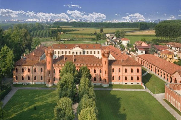 Historická tehlová budova s vežami uprostred zelenej trávy a stromov, s horami v pozadí.