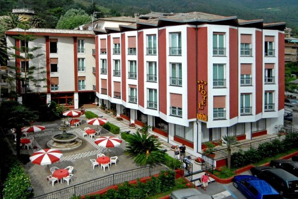 Hotel s červenobielymi slnečníkmi na terase, obklopený zeleňou a horami.