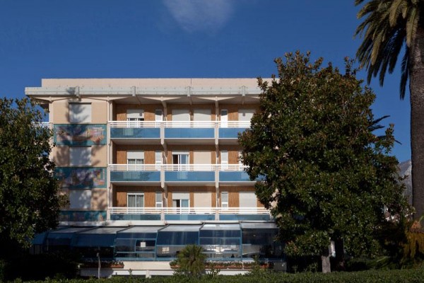 Štvorposchodová budova s balkónmi, obklopená stromami a modrou oblohou.