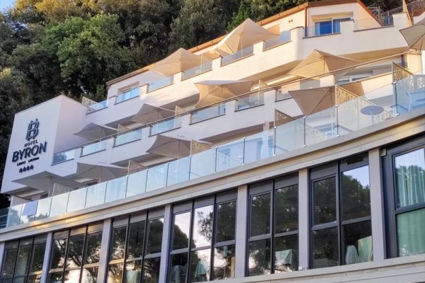 Moderný hotel so sklenenými balkónmi a slnečníkmi zasadený v zeleni.