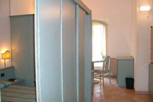 Jednoducho zariadená hotelová izba s posteľou, lampou a menším stolom pri okne.