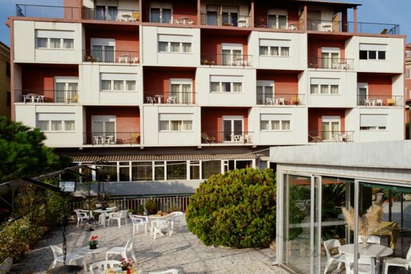 Svetlá budova s tromi poschodiami balkónov nad terasou s bielym vonkajším nábytkom.