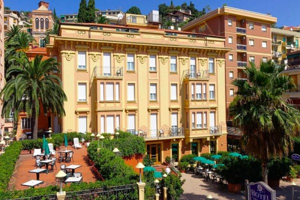 Žltý hotel s terasou a palmami na pozadí mesta.