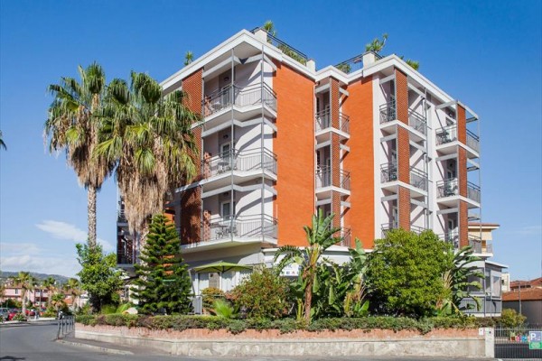 Moderná štvorposchodová budova s balkónmi obklopená palmami a zeleňou.