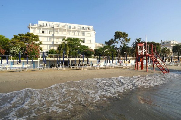 Pláž s ležadlami, slnečníkmi a strážnou vežou, v pozadí hotel.