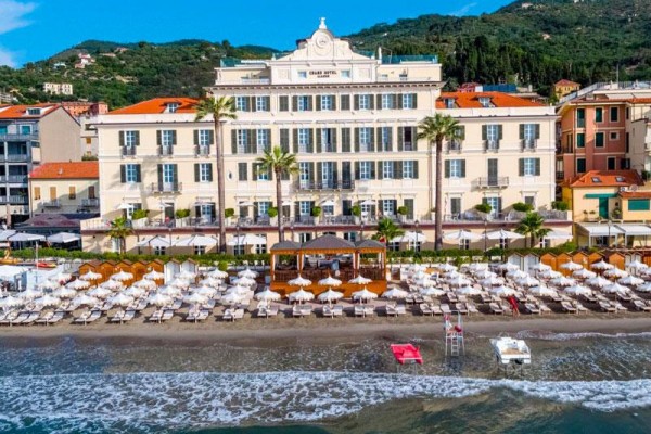 Grand hotel pri pláži s lehátkami a slnečníkmi, obklopený palmami.