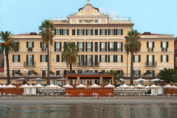 Veľký hotel na pláži s palmami a terasou s bielymi slnečníkmi.