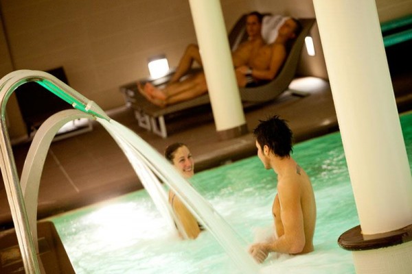 Dvaja ľudia relaxujú v bazéne s vodopádom, zatiaľ čo dvaja ďalší odpočívajú na ležadlách v pozadí.